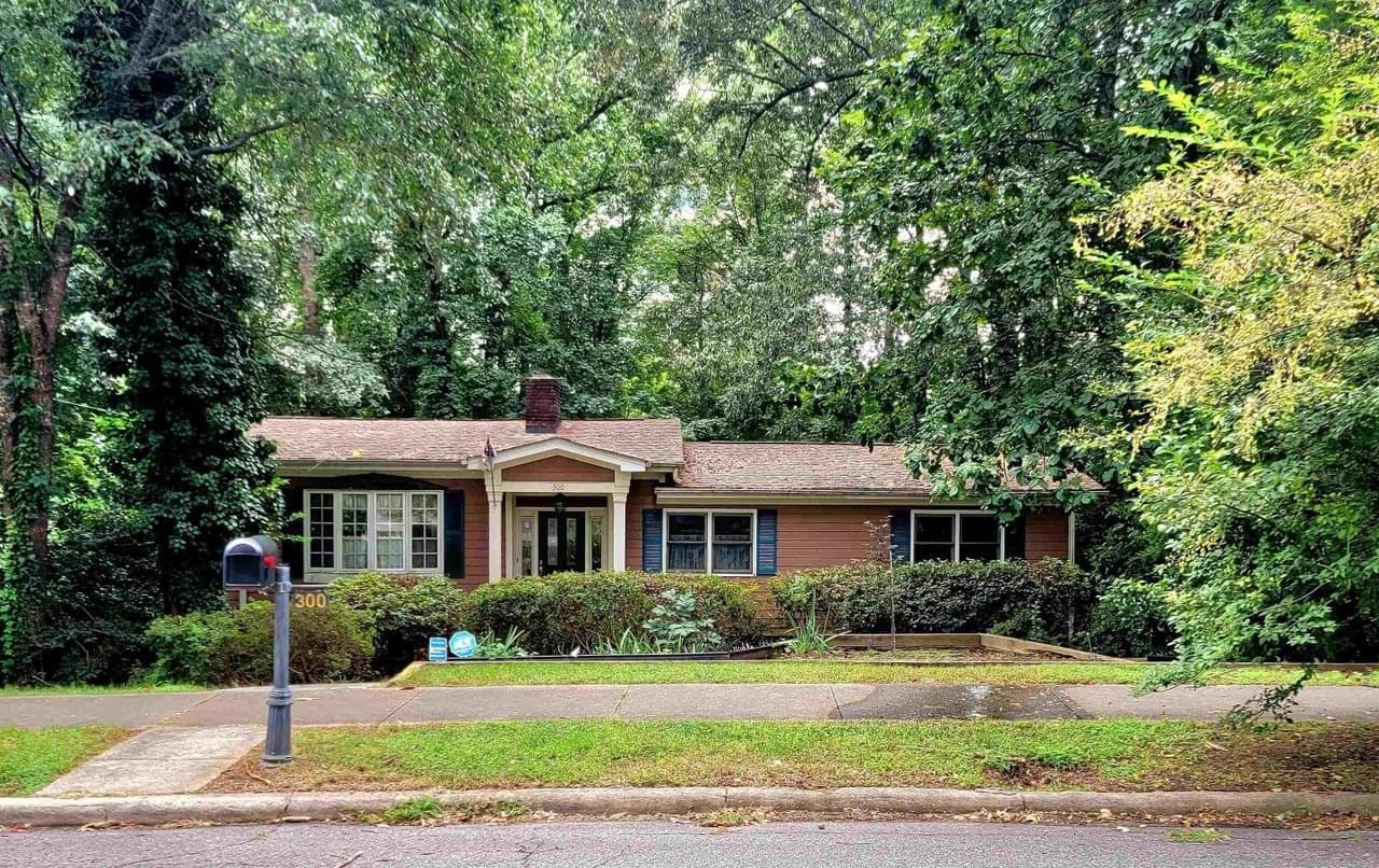 A single-story home with red-brown siding, blue shutters, and white trim on a lush forested lot